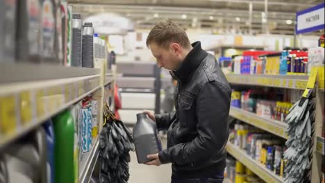 Young-man-is-choosing-motor-oil-for-car-at-repair-service-station-or-in-supermarket.-He-looks-on-a-few-canisters,-chooses-one-and-goes-to-pay-for-it.-Different-canisters-on-shelves