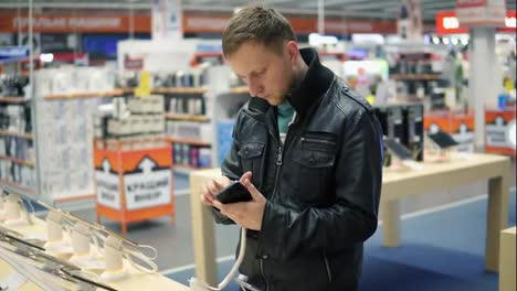 Young-man-in-black-leather-jacket-is-choosing-a-new-mobile-phone-in-a-shop,-checking-how-it-works