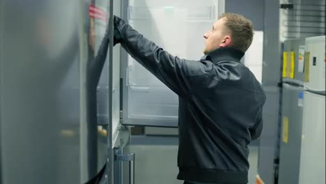 Young-man-is-choosing-a-refrigerator-in-a-store.-He-is-opening-the-doors,-looking-inside