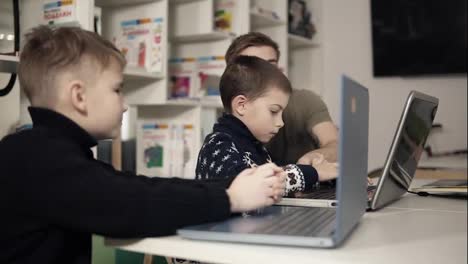 Dos-Niños-Pequeños-Están-Aprendiendo-A-Usar-Computadoras-Portátiles-Con-La-Ayuda-Y-Orientación-De-Un-Joven-Maestro-Programador-Sentado-A-Su-Lado-En-Un-Salón-De-Clases.