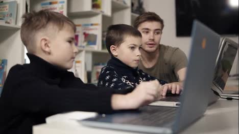 Young-attractive-male-teacher-showing-two-little-boys-how-to-use-gadgets-such-as-laptop.-Technologies-and-progress.