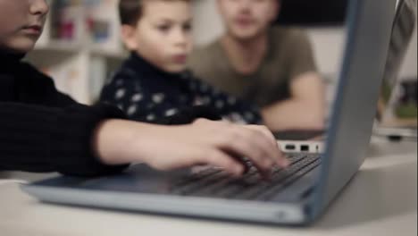 Kid's-arms-typing-somethingon-laptop-keyboard-with-two-unrecognizable-figures-sitting-on-the-background.-Programmingfor-kids-class.-Learning-how-to-use-technologies-and-gadgets.