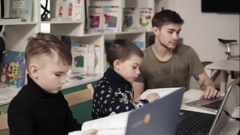 Two-little-boys-are-reading-books-related-to-computers-and-programming.-Educational-process.-Young-attractive-male-teacher-is-explaining-them-how-gadgets-work.
