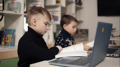 Two-little-boys-reading-books-in-a-classroom-sitting-in-front-of-laptops.-Educational-process.