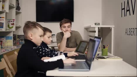 Young-attractive-male-teacher-in-programming-class-helps-two-little-boys-learn-something-about-gadgets-such-as-laptops.-Educational-process.