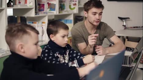 Attarctive-male-programmer-teacher-with-several-tattooes-on-his-arms-explaining-something-to-his-two-little-pupils-boys-on-a-programming-class.-Alternative-education-for-children.