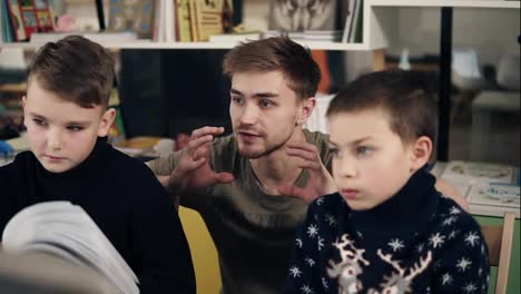 Attractive-young-male-teacher-with-couple-tattooes-on-his-arms-explaining-something-to-two-little-boys-in-warm-cozy-sweaters.
