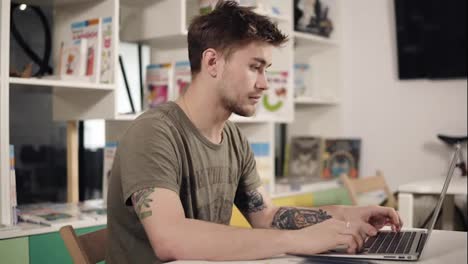 Attractive-young-male-programmer-teacher-with-several-tattooes-on-his-arms-is-mumbling-something-while-typing-on-laptop-keyboard.