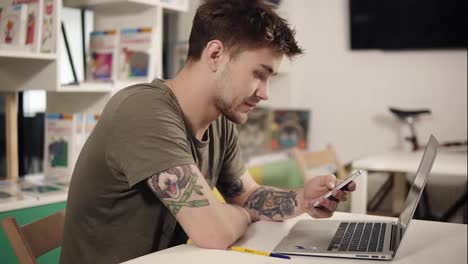 Attractive-programmer-with-tattooes-on-his-arms-scrolling-something-on-his-smartphone-while-sitting-in-a-classroom-beside-laptop.