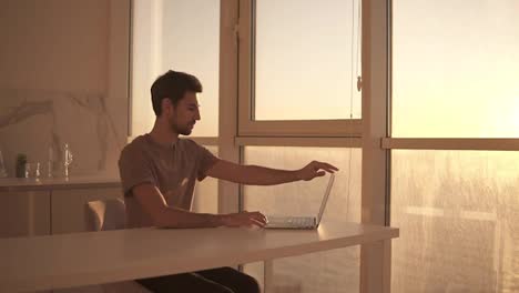 European-dark-haired-man,-freelancer-opens-laptop-for-work.-Young-guy-sitting-close-to-panoramic-window-on-studio-kitchen---focused-on-working-with-laptop-and-making-difficult-task.-Slow-motion