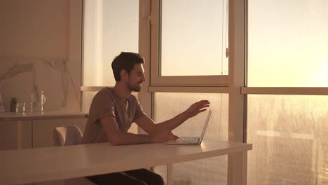European-dark-haired-man-sitting-close-to-panoramic-window-on-studio-kitchen---looking-on-laptop-deskop-and-cheerfully-smiling,-chatting-with-friends.-Slow-motion.-Side-view