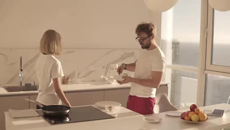 Relaxed-caucasian-man-preparing-breakfast-for-two-in-kitchen-at-home-and-mixing-eggs-in-bowl,-blonde-girl-preparing-spices.-Happy-couple-in-white-T-shirts-smiling-and-chatting.-Studio-kithen.-Sunlight