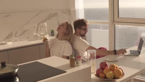 Side-view-of-bearded-man-working-on-laptop-computer-sitting-at-home-on-a-kitchen-with-panoramic-windows.-Young-blonde-wife-sitting-from-behind-and-enjoying-eating-her-apple-in-the-morning