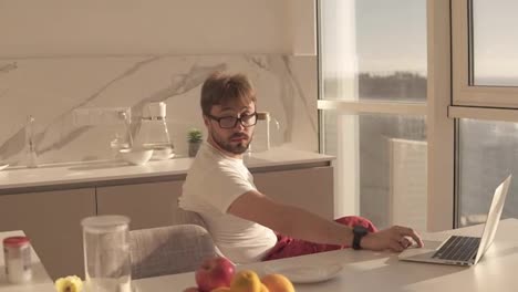 Bearded-man-in-eyes-glasses-working-on-laptop-computer-sitting-at-home-on-a-kitchen-with-panoramic-windows.-Young-wife-embracing-him-from-behind-and-they-kissing-each-other