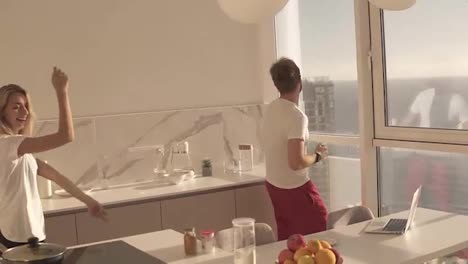 Young-caucasian-couple-wearing-pyjamas-funnily-dancing-and-singing-early-in-morning,-preparing-for-new-day,-spending-time-together,-having-fun.-Studo-kitchen-with-big-windows-on-the-background
