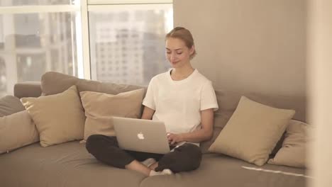 Smiling-woman-freelancer-sitting-on-couch-in-domestic-clothes,-freelance-from-home,-typing-email-on-laptop.-Focused-blonde-girl-using-computer-while-sitting-relaxfully-on-a-couch
