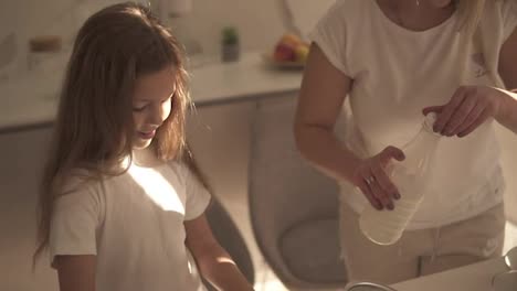 Mother-helping-and-teaching-her-cute-daughter-in-modern-cozy-white-kitchen-to-cook-cake-or-pancakes.-Girl-adding-milk-and-mixing-ingredients.-Happy-family.-Relationship-mom-and-daughter