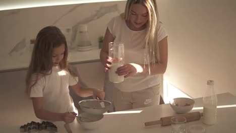 Mother-helping-and-teaching-her-cute-daughter-in-modern-cozy-white-kitchen-to-cook-cake-or-pancakes.-Mother-adding-flour-through-a-sieve-and-girl-mixing-ingredients.-Happy-family.-Relationship-mom-and-daughter