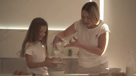 Mother-helping-and-teaching-her-cute-daughter-in-modern-cozy-white-kitchen-to-cook-cake-or-pancakes.-Mother-adding-flour-through-a-sieve-and-girl-mixing-ingredients.-Having-fun-with-flour