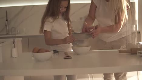 Mother-helping-and-teaching-her-cute-daughter-in-modern-cozy-white-kitchen-to-cook-cake-or-pancakes.-Mother-adding-flour-through-a-sieve-and-mixing-ingredients.-Having-fun-with-flour.-Slow-motion