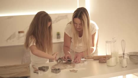 Mother-and-daughter-both-with-long-hair-cut-out-different-cookie-shapes-from-the-dough-in-kitchen.-Mom-and-daughter-cooking-christmas-cookies-at-home-and-having-fun