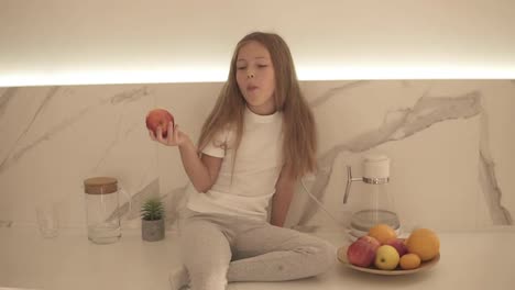 Little-girl-with-long-fair-hair-eating-red-apple-in-bright,-modern-kitchen-while-sitting-on-a-counter.-Bowl-of-fresh-fruits-next-to-her.-Girl-enjoying-fresh-apple