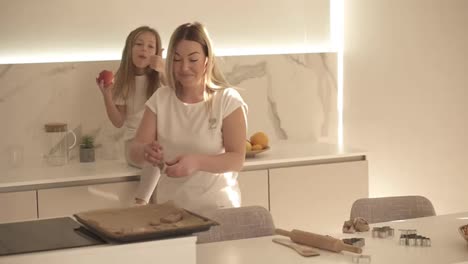 Laughing-mom-and-daughter-having-fun-togethe.-Girl-is-sitting-on-a-kitchen-counter,-eating-apple-while-mom-is-arranging-cookies-on-tray.-Mother-kissing-her-daughter.-Slow-motion