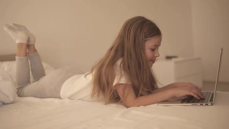 Long-haired-european-girl-typing-on-laptop.-Smiling-little-girl-with-interest-watch-laptop-and-lay-on-the-bed.-Side-view,-slow-motion