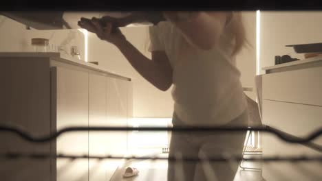View-from-the-oven:-long-haired-teen-girl-opens-the-oven's-door-and-puts-a-baking-pan-inside.-Beautiful-girl-on-kitchen-puts-her-selfmade-cookies-into-the-oven