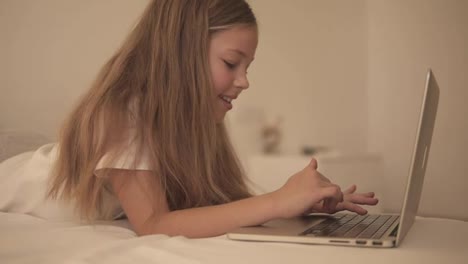 Close-up-of-long-haired-beautiful-european-girl-typing-on-laptop.-Smiling-little-girl-with-interest-watch-laptop-and-lay-on-the-bed.-Side-view,-slow-motion