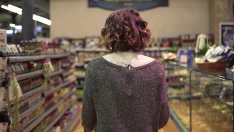 Rare-view-of-a-woman-with-wavy-hair-is-driving-shopping-trolley-through-food-department-in-supermarket-and-looking-around.-Pretty-girls,-everyday-life-and-buying-products-concept