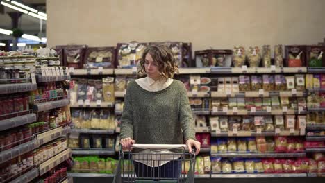 Portrait-of-a-woman-with-wavy-hair,-wearing-casual-is-driving-shopping-trolley-through-the-row-withfull-of-goods-shelf-in-supermarket-and-looking-around.-Pretty-girls,-everyday-life-and-buying-products-concept