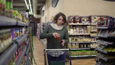 Front-view-of-caucasian-woman-walk-with-a-cart-near-shop-shelves-choosing-a-glass-jar-in-grocery-market-and-put-it-to-the-cart.-Female-customer-checking-product-ingredients.-Supermarket,-sale,-shopping,-assortment,-consumerism-concept
