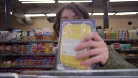 Young-curly-haired-woman-doing-grocery-shopping-at-the-supermarket,-she-is-reading-a-product-label-and-nutrition-facts-on-a-box-with-cheese.-Footage-from-the-shelf