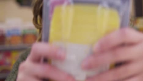 Young-curly-haired-woman-doing-grocery-shopping-at-the-supermarket,-she-is-reading-a-product-label-and-nutrition-facts-on-a-box-with-cheese.-Footage-from-the-shelf,-exremely-closeup-of-face