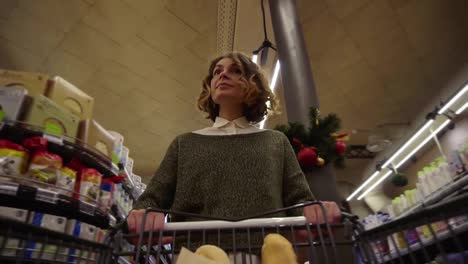 Pretty-woman-in-casual-clothes-is-walking-in-grocery-store-steering-shopping-trolley-with-bread-inside-it-and-looking-around-at-shelves-with-products.-Women-and-shops-concept.-Low-angle-footage-from-the-cart