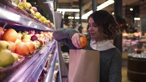 Cute-girl-buys-fresh-oranges-in-the-market.-Beautiful-young-woman-stands-in-front-the-shelf-and-puts-the-oranges-to-a-brown-paper-bag,-she-is-pleased-with-the-choice