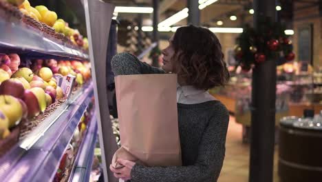 Cute-girl-buys-fresh-red-apples-in-the-market.-Beautiful-young-woman-stands-in-front-the-shelf-and-puts-the-apples-to-a-brown-paper-bag,-she-is-pleased-with-the-choice.-Slow-motion