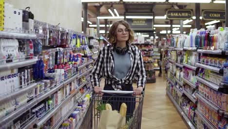 Modern-young-girl-in-glasses-and-headphones-on-neck-walking-with-shopping-cart-in-the-supermarket-and-stearing-around.-Girl-in-plaid-shirt-and-jeans.-Slow-motion