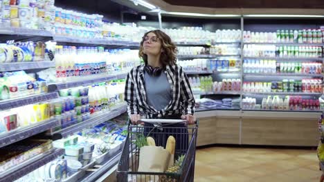 Modern-young-carefree-girl-in-glasses-and-headphones-on-neck-walking-with-shopping-cart-in-the-supermarket-and-stearing-around.-Girl-in-plaid-shirt-and-jeans-in-a-happy-mood.-Slow-motion