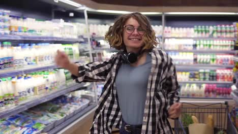 At-the-supermarket:-happy-young-girl-funny-dancing-between-shelves-in-supermarket.-Curly-girl-wearing-jeans-and-black-and-white-plaid-shirt-with-headphones-on-neck.-Slow-motion