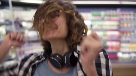 At-the-supermarket:-happy-young-girl-funny-dancing-between-shelves-in-supermarket.-Curly-girl-wearing-black-and-white-plaid-shirt-with-headphones-on-neck.-Slow-motion.-Close-up