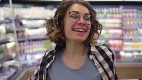 At-the-supermarket:-happy-young-girl-funny-dancing-between-shelves-in-supermarket.-Curly-girl-wearing-black-and-white-plaid-shirt-with-headphones-on-head-listening-to-her-favorite-music.-Slow-motion.-Close-up