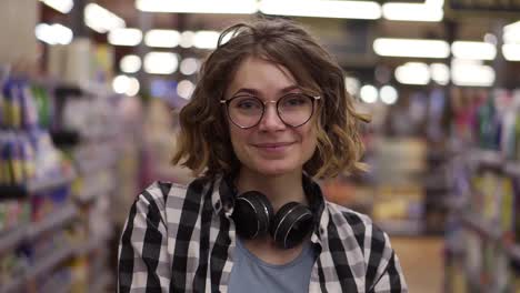 Portrait-young-woman-stands-in-front-of-the-camera-and-smiles-in-supermarket-feel-happy-girl-shopping-face-retail-store.-Pretty-curly-female-customer-in-casual-with-headphones-on-neck