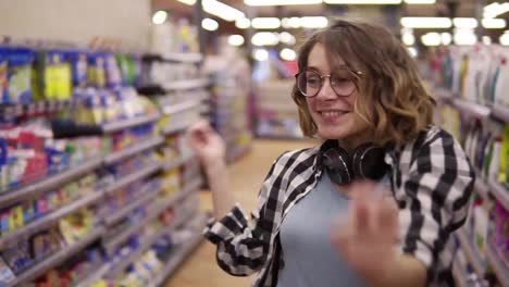 Close-up-portrait-of-young-woman-in-plaid-shirt-and-glasses-dancing-standing-at-grocery-store-aisle.-Excited,-smiling-with-headphones-on-neck-woman-having-fun,-dancing-at-supermarket.-Slow-motion