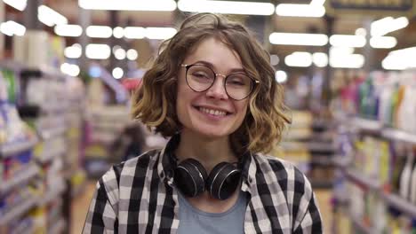 Portrait-young-woman-stands-in-front-of-the-camera-and-smiles-in-supermarket-feel-happy-girl-shopping-face-retail-store.-Pretty-curly-female-customer-in-casual-with-headphones-on-neck-with-other-customers-on-blurred-background