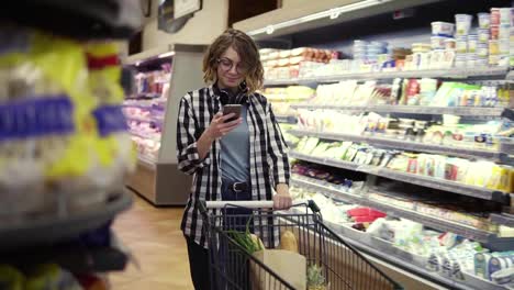 Shopping,-technology,-sale,-consumerism-and-people-concept---woman-with-smartphone-and-headphones-on-neck-walking-at-supermarket.-Smiling-woman-in-glasses-texting-with-friends,-walks-with-her-shopping-trolley.-Slow-motion