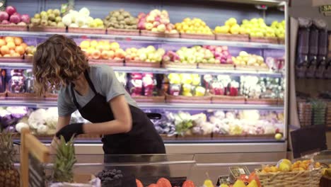 Portrait-of-female-shop-assistant-in-gloves-and-black-apron-filling-up-fruits-and-vegetables-storage-stands-with-assorted-organic-groceries-in-supermarket.-Slow-motion