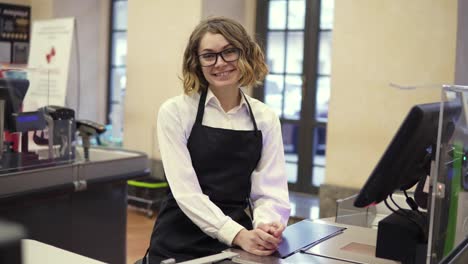 Woman-in-black-apron-as-a-cashier-at-the-cash-register-in-the-supermarket-or-discounter.-Smiling-short-haired-curly-woman-smiling-to-the-camera