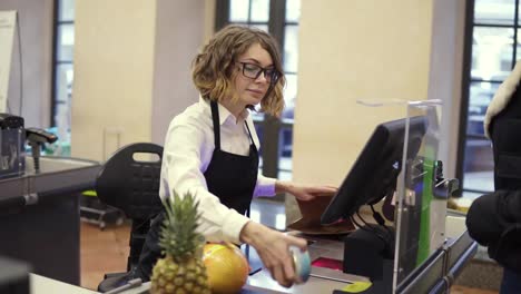 Cheerful-slender-saleswoman-in-white-shirt-and-black-apron-scanning-product,-fruits-at-checkout-counter-in-bright-supermarket-and-putting-it-into-brown-paper-bag-while-unrecognizable-customer-is-waiting